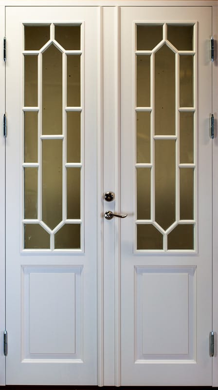 En vit dubbeldörr med glasinfattningar som täcker den övre två tredjedelarna av varje dörrblad. Glasrutorna är indelade i geometriska former med dekorativa spröjsar som bildar rektanglar och spetsiga bågar. Den nedre tredjedelen av dörrbladen har rektangulära speglar. Dörren är utrustad med metallhandtag och lås placerade centralt.