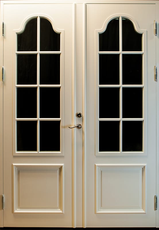 En vit dubbeldörr med eleganta glaspartier. Varje dörrblad har ett högt rektangulärt glas med spröjs som delar in ytan i sex mindre rutor. Glaspartiernas överdel avslutas med en dekorativ bågform. Nederdelen av dörrarna pryds av en rektangulär panel. Handtaget och dörrlåset är placerade i mitten.