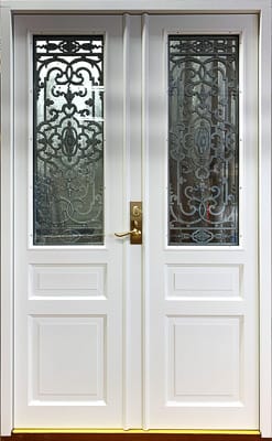 Vit dubbeldörr med klassisk design. Varje dörrblad har en stor glasruta i övre delen, dekorerad med ornamentalt smidesmönster i svart. Nederdelen av dörrbladen har två rektangulära speglar. Dörren har ett gyllene handtag på höger sida.
