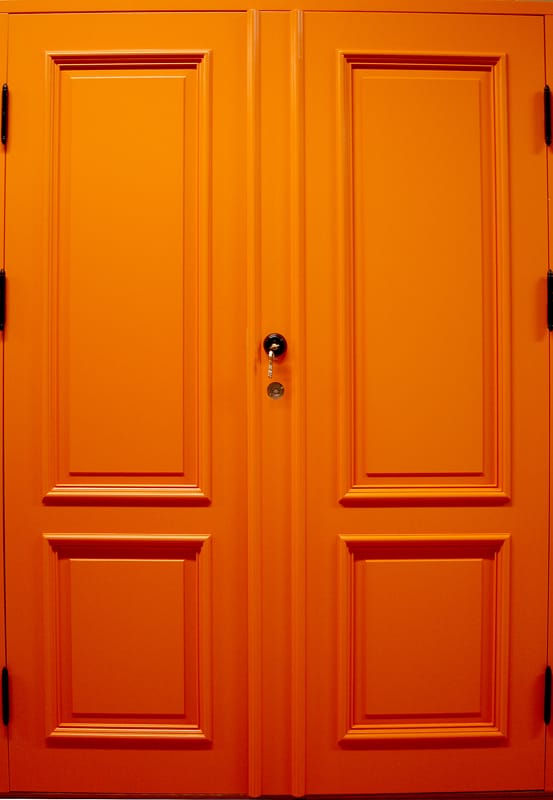 Klargul-orange dubbeldörr i klassisk design utan glasrutor. Dörrarna har fyra rektangulära spegelpaneler med profilerade kanter. Handtaget är runt och placerat mitt på vänster dörr.