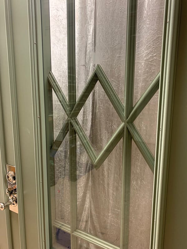 Närbild på en grön dubbeldörr tagen snett från sidan. Dörren har en glasruta med frostat glas och dekorativa spröjsar i diamantmönster. I hörnen finns små blå glasdetaljer. Nederdelen av dörren har en panel med en upphöjd rombform. Ett silverfärgat handtag syns till vänster.
