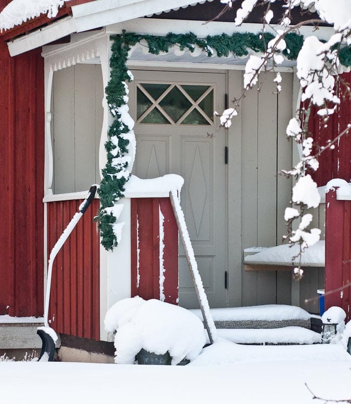 Grå ytterdörr med krysspröjsat överljus på en snötäckt farstukvist till en röd trästuga.