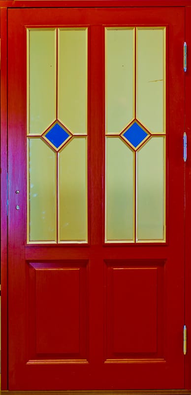 Röd ytterdörr med två höga glaspartier i varmt gultonat glas, vardera med en blå rombformad detalj i mitten, samt två rektangulära speglar i nedre delen och gångjärn på höger sida.