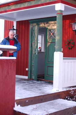 Röd träfasad med grön ytterdörr vintertid och man i blå jacka på snötäckt farstukvist