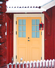 Gul pardörr med glasrutor upptill på ett rött trähus, omgiven av snö och med en röd lykta hängande vid sidan.