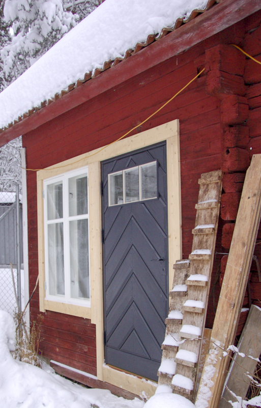 En mörkgrå ytterdörr med fiskbensmönster och tre glasrutor i överkant sitter i en röd timrad byggnad med ljus träram runt dörr och fönster. Snö täcker marken, taket och en lutande träläktare som står mot väggen.