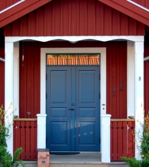 Blå dubbeldörr med klassisk paneldesign, placerad i en röd träfasad med vit inramning och veranda.