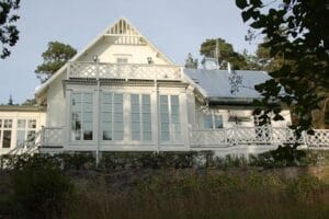 Verandafasad på vit trävilla med höga spröjsade fönsterpartier och glasdörrar samt balkong med vit krysspanel.