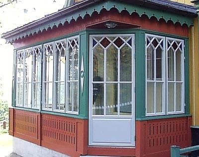 Inglasad veranda med vit pardörr och fönster med dekorativt spröjsmönster, omgiven av grön och röd träpanel.