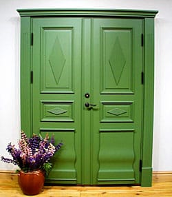 Foto av grön allmogeinspirerad pardörr i trä med profilerade spegelpaneler och diamantformade dekorrutor, omgiven av kraftig dörrkarm och med en kruka med lila blommor placerad på golvet framför.