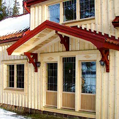 Ett hus med ljus träfasad och mörkrött sadeltak. Vid entrén finns en farstukvist med rött snedtak och dekorativa trästöd. Fasaden har stora spröjsade fönster och glasade dörrar med trärutor i nederkant. Två svarta vägglampor sitter på varsin sida om entrén. Snö ligger på marken och på taket.