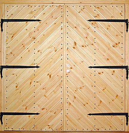 Pardörr i ljust trä med tät diagonal panel i V-mönster. Dörren är förstärkt med dekorativa smidda svarta gångjärn, tre på varje dörrblad. Små svarta spikar sitter jämnt placerade längs panelen och skapar en markerad ram runt hela dörren.