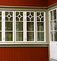 Närbild på vita spröjsade fönster i rad på en glasveranda. Fönstren har dekorativa mönster med kvadratiska spröjs i övre delen. Fasaden är målad i rött med grågrön inramning kring fönstren och vitmålad dörr till höger.