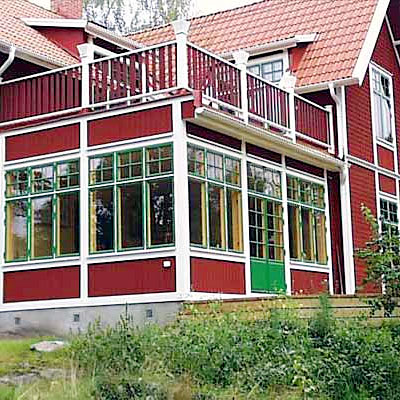 Tvåvånings rött trähus med vita knutar och gröna fönsterkarmar. Bottenvåningen har en stor glasveranda med spröjsade fönster och en grönmålad dörr. Ovanför finns en balkong med vitt räcke. Huset har rött tegeltak.