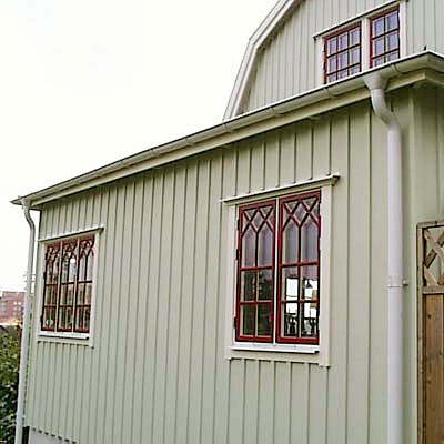 Husfasad i ljusgrön träpanel med flera fönster som har brunröda karmar och dekorativ spröjsning i spetsbågsmönster. Huset har vit hängränna och stuprör, samt en träport till höger i bilden.