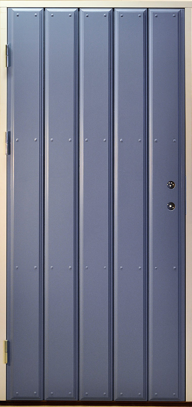 Enkel ytterdörr i blå kulör med stående panel och markerade spår mellan varje bräda. Dörren är dekorerad med små runda nitar längs panelerna, vilket förstärker det traditionella uttrycket. På höger sida sitter två blanka metallbeslag för lås och handtag. Dörrens enkla och rena design ger ett tidlöst och klassiskt intryck.