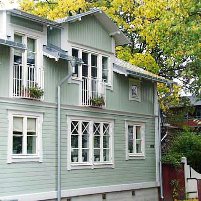 Tvåvåningshus i ljusgrön träpanel med vita detaljer. På nedervåningen syns ett vitt tvådelat fönster med krysspröjs och små krukor i fönsterkarmen. På övervåningen finns höga smala fönster med vita räcken och blomlådor med gröna växter. Taket har vitmålade takutsprång och runt huset finns träd med gröna och gula löv.