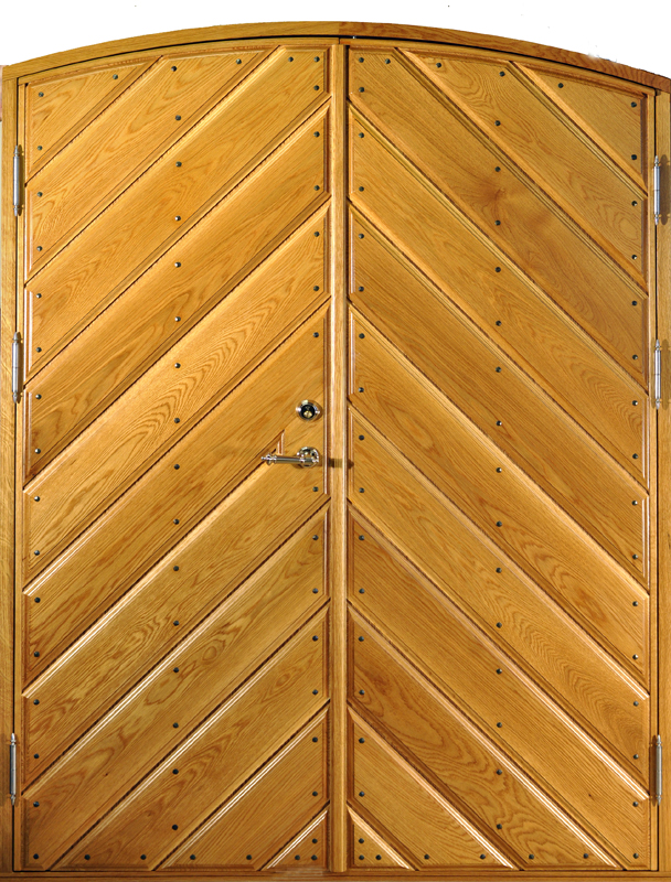 Pardörr i ek med rundad överkant och diagonal paneldesign. Panelerna är lagda i ett fiskbensmönster med metallnitar längs varje bräda, vilket ger ett robust och dekorativt utseende. Dörren har en varm, gyllene träton med tydlig ådring. På vänstra dörrbladet sitter ett blankt metallhandtag och en rund låscylinder, vilket förstärker den klassiska och gedigna känslan.