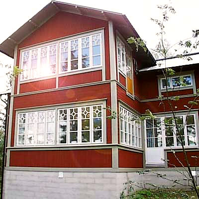 Tvåvåningshus i klassisk stil med röd träfasad och grå sockel. Fasaden pryds av stora vita fönster med spröjs i dekorativa mönster, där vissa rutor är formade som åttahörningar. På nedervåningen syns en vit dörr med glaspartier, och ovanför finns en glasveranda som ger ett luftigt intryck. Huset har ett sadeltak och omges av träd och grönska.