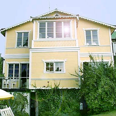 Ett tvåvåningshus i klassisk stil med gul träfasad och vita snickeridetaljer. Huset har flera spröjsade fönster i olika storlekar, bland annat ett burspråk på övervåningen med sex rutor. På nedervåningen finns en balkongdörr med spröjs samt ett mindre fyrdelat fönster i mitten. Taket har vita snickarglädje-detaljer längs takfoten. Solens reflex syns i ett av övervåningens fönster. Gröna klängväxter växer upp längs fasaden.