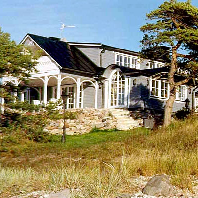 Stort hus i två våningar målat i grått med vita detaljer. Huset har en generös veranda med vita träpelare och bågar, samt stora fönsterpartier med spröjs. En stentrappa leder upp från trädgården till entrén. Taket är svart och brant, och huset omges av tallar och naturlig vegetation.