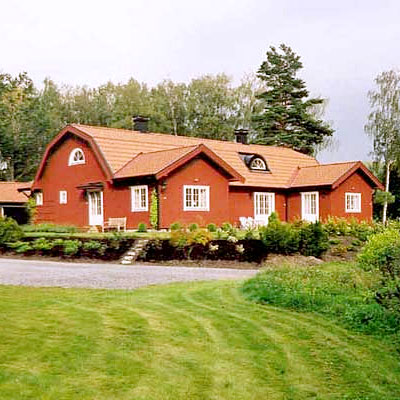 Traditionell svensk villa målad i falurött med ljusa foder runt fönstren. Huset har tegeltak i varm orangeröd ton och flera takkupor samt små runda fönster i gavlarna. Trädgården är välskött med gröna buskar längs fasaden, en grusgång och klippt gräsmatta i förgrunden. I bakgrunden syns skog med höga björkar och granar.