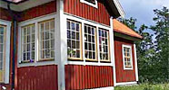 En röd träbyggnad med vita knutar och spröjsade fönster. Fönsterpartiet är stort och omfattar flera sektioner, vilket skapar ett ljust och öppet intryck. Byggnadsstilen är klassiskt svensk och påminner om äldre torp och villor på landsbygden.