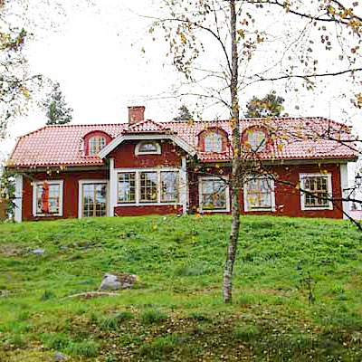 En stor röd villa i klassisk svensk stil med vita knutar och spröjsade fönster. Huset har rött tegeltak med flera takkupor som ger en charmig karaktär. På bottenvåningen finns en inglasad veranda med stora fönster, och huset är omgivet av grön gräsmatta och träd.