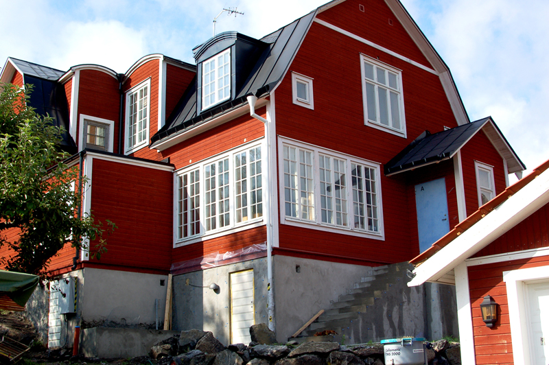 Ett stort rött trähus med vita foder runt spröjsade fönster och svart plåttak. Huset har flera burspråk och takkupor med fönster samt en glasveranda som löper längs ena sidan. Sockeln är i betong och det pågår byggnation vid grunden, med synliga stenar, verktyg och en ofärdig trappa upp mot entrén. I förgrunden syns delar av ett mindre rött uthus med vitmålad takkantslist.