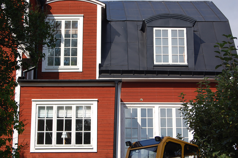 Rött trähus med svart plåttak och flera vita spröjsade fönster i olika storlekar. På bottenvåningen syns ett större fyrdelat fönster och ett tvådelat fönster, medan övervåningen har två höga fönster med många små rutor – ett på en frontespis och ett i en takkupol. Framför huset står en gul maskin och grenar från träd skymmer delvis fasaden.