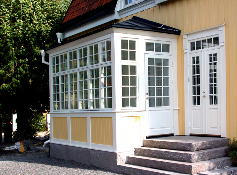 En gulmålad villa med glasveranda och vita spröjsade fönster. Verandan har vitmålade partier nedtill och en glasad dörr med spröjs. Bredvid verandan finns en vit dubbeldörr med glasrutor och spröjs, som leder in i huset. Trappsteg i sten leder upp till båda ingångarna, och i bakgrunden syns grönska.
