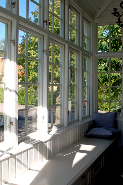Ett ljust uterum eller en glasveranda med stora vitmålade spröjsade fönster som släpper in mycket dagsljus. Längs fönsterväggen finns en vitmålad sittbänk med kuddar i blått, vilket skapar en inbjudande och avkopplande plats. Utanför syns grönska och träd i solsken.