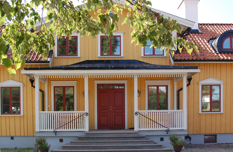 Gulmålad trävilla med vit veranda och röd entrédörr i mitten. Huset har röda fönsterbågar med vita omfattningar och dekorativa triangulära överstycken ovan vissa fönster. Röda takpannor pryder taket, och ett burspråk med svartkupat fönster syns på höger sida. Gröna lövträd ramas in i förgrunden.