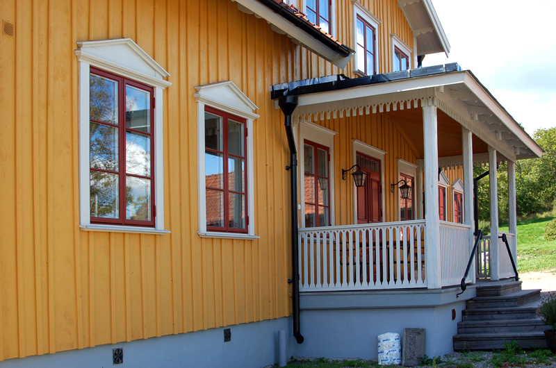 En gulmålad träfasad med stående panel och rödmålade fönster indelade i fyra rutor. Fönstren har vita omfattningar med triangulära överstycken. Till höger syns en vitmålad veranda med snickarglädje i räcket och under takfoten. Verandan har en trappa i trä som leder ner till marken och bredvid sitter två svarta vägglyktor.