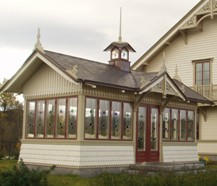 En ljus träveranda med dekorativa detaljer i klassisk stil, kopplad till ett större hus. Byggnaden har många små fönster med färgade glasrutor i övre delen, röd dörr och röda fönsterbågar. Taket är mörkt och kröns av en liten spira och tornliknande detalj, vilket ger ett utsmyckat och traditionellt utseende.