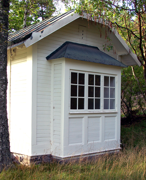 Litet vitt trähus eller uthus med sadeltak i mörk plåt. På framsidan sitter en sektion med spröjsade fönster och nedre panel utan glas, liknande en veranda i miniatyr. Huset står på en låg grund av sten och omges av högt gräs och träd i bakgrunden. Fasaden är målad i vitt med liggande panel.