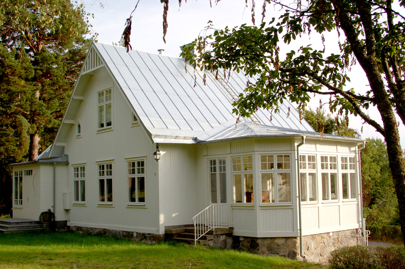 Vitt trähus i klassisk stil med högt sadeltak i silvergrå plåt. På gaveln syns en inglasad veranda med spröjsade fönster åt två håll, placerad på en grund av natursten. Huset har symmetriskt placerade fönster med vita ramar, en enkel trappa med smidesräcke upp till verandan och en gammaldags vägglykta bredvid. I förgrunden finns en grön gräsmatta och ett träd med hängande grenar som ramar in bilden.