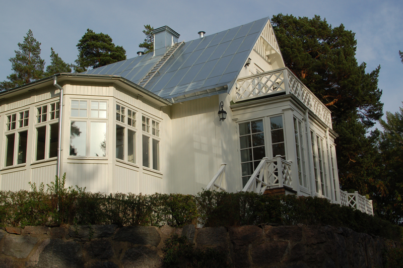 Vitt trähus i klassisk stil med högt sadeltak i silvergrå plåt. Huset står på en murad stenfot och har en vinklad glasveranda med stora spröjsade fönster åt två håll. Till höger syns en upphöjd altan med vita kryssräcken och helglasade pardörrar. Taket har en brant lutning och är utrustat med en stege för sotaraccess. Omkring huset växer täta buskar och bakom reser sig stora tallar mot himlen.