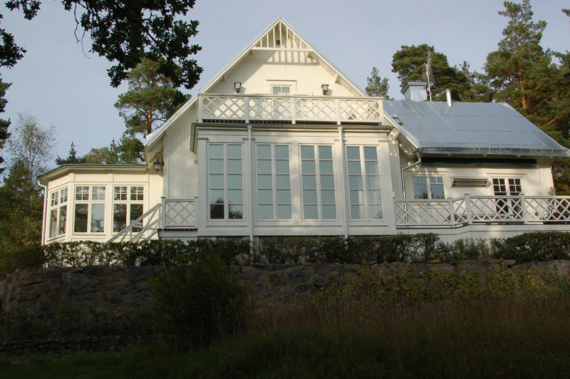 Stort vitt trähus i klassisk stil med sadeltak i silvergrå plåt. Fasaden domineras av en glasveranda med helglasade dubbeldörrar och spröjsade fönster, som sträcker sig nästan hela höjden från mark till tak. Ovanför finns en bred balkong med vitmålat kryssräcke. Till vänster syns en vinklad glasveranda med stora fönster, och till höger ytterligare en altan med samma typ av kryssräcke. Huset är upphöjt på en stenmur och omges av träd och buskar.