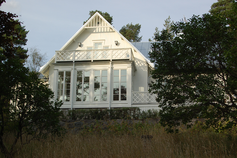 Vitmålad trävilla i två våningar med sadeltak i silvergrå plåt. Huset har en stor glasveranda med höga helglasade dörrar och spröjsade fönster från golv till tak, vilket ger ett luftigt intryck. Ovanför verandan finns en balkong med vitmålat kryssräcke. Huset ligger på en höjd omgiven av natur med buskar, träd och högt gräs i förgrunden.