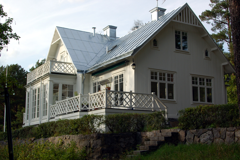 Tvåplansvilla i klassisk stil med vit träfasad och silvergrått plåttak. Huset har flera spröjsade fönster i olika storlekar samt en inglasad veranda med helglasade dörrar och fönsterpartier. På framsidan finns en altan med vitmålat kryssräcke, och ovanför en balkong i samma stil. Huset är byggt på en liten höjd med stenmurar och trappsteg som leder upp från trädgården, omgiven av buskar och höga träd.