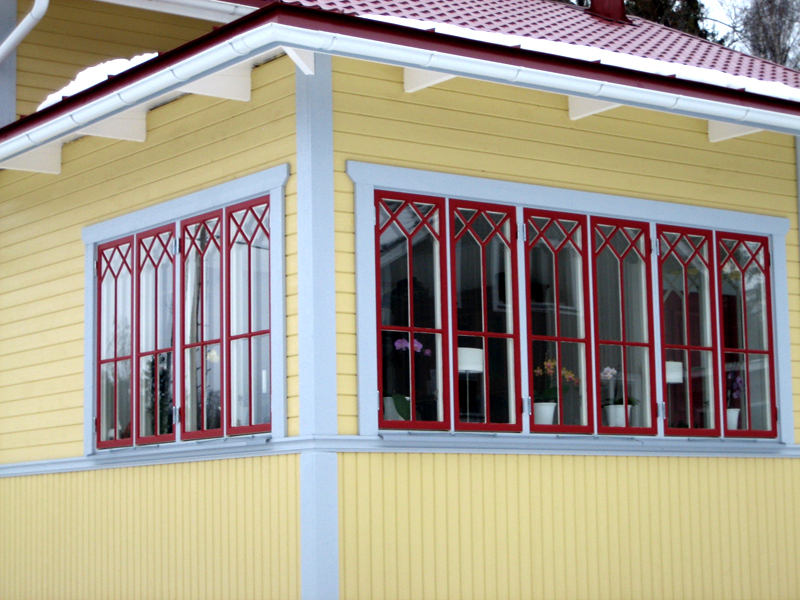 Närbild av ett gult trähus med röda spröjsade fönster och ljusgrå foder. Fönstren har dekorativa krysspröjs upptill och står i två rader längs en hörnveranda. Inne i huset syns krukväxter med blommor placerade i fönstren. Taket är rött och täckt av snö, medan fasaden har både liggande och stående panel.