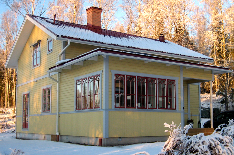 Tvåvåningshus med gul träfasad, gråa snickerier och rött sadeltak täckt av snö. Huset har många fönster med röda karmar, varav de stora på verandan är dekorerade med spröjs i mönster. En skorsten i tegel sticker upp från taket. Marken och träden runt huset är täckta av snö, vilket skapar en vintermiljö. I bakgrunden syns solbelysta träd.