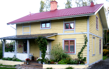 Tvåvåningshus med gul träfasad, vitt och grått snickeri samt rött sadeltak. Huset har flera fönster med röda karmar och en liten veranda med vitt räcke och växter runtomkring. En klätterväxt växer upp längs fasaden och en spaljé sitter vid ena väggen. I förgrunden syns en trädgård med buskar, blommor och en stenbelagd gång. Bakom huset skymtar träd.