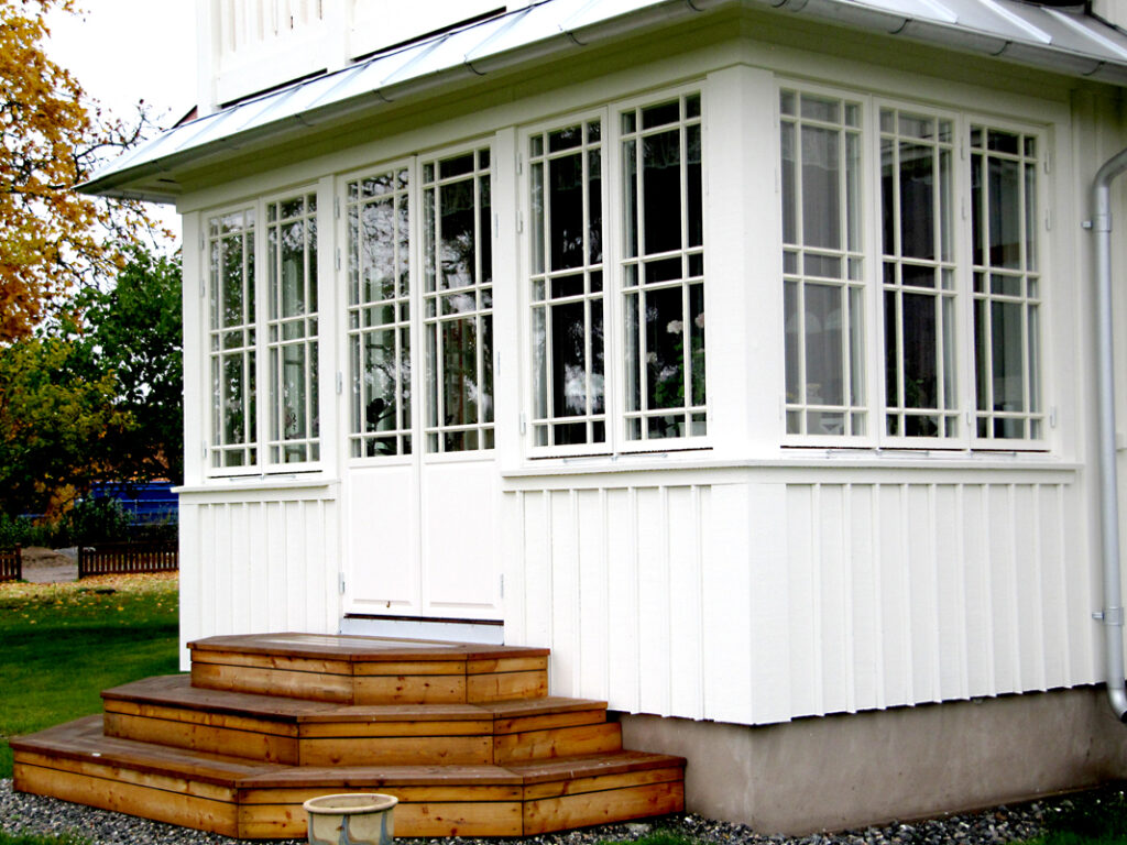Närbild på en vitmålad inglasad veranda på en villa. Verandan har stora spröjsade fönster som går runt hela hörnet och en vit dubbeldörr i mitten. Nedanför dörren leder en bred, terrassformad trappa i trä ner mot gräsmattan. Grus ligger längs husgrunden, och i bakgrunden syns höstfärgade träd och en liten trästaket.
