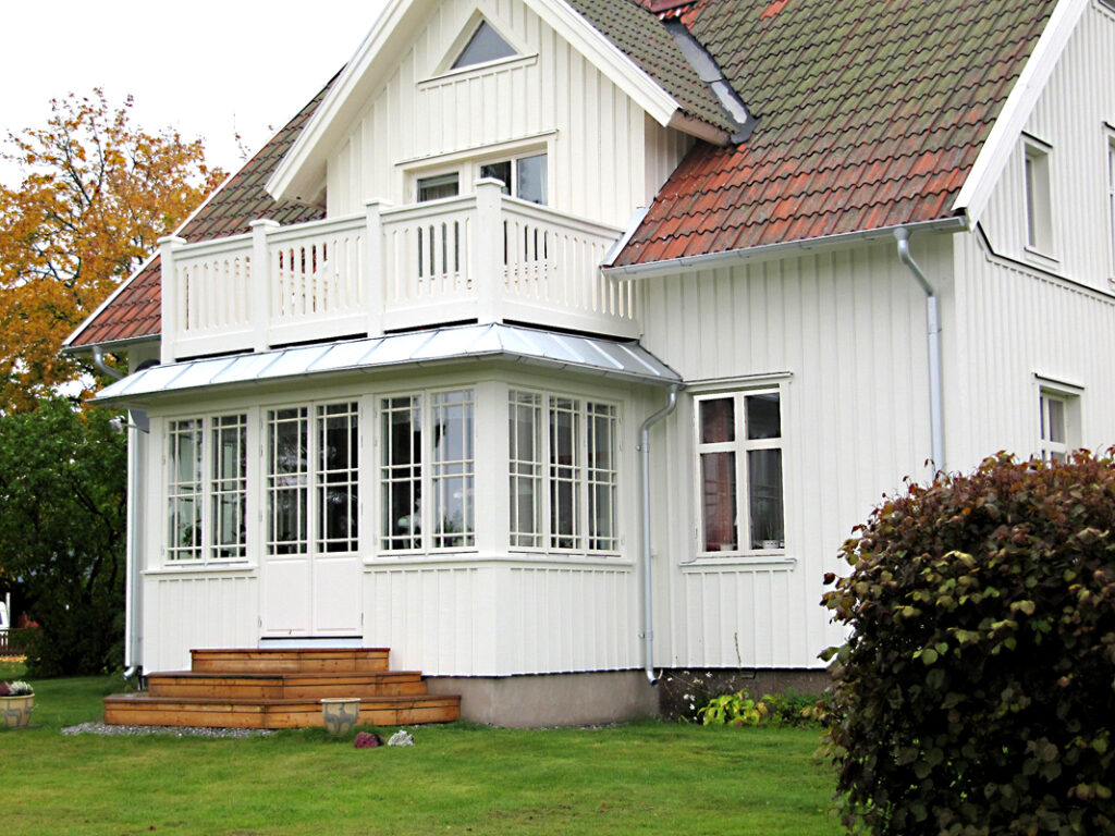 En vit träpanelklädd villa med sadeltak i tegel, där en inglasad veranda med stora spröjsade fönster och dubbeldörrar byggts till. Verandan har ett plåttak och leder ut mot en liten trappa i trä som mynnar i en välklippt gräsmatta. På övervåningen finns en balkong med vita räcken. Buskar och träd runt huset ger en grönskande inramning, och ett höstfärgat träd syns i bakgrunden.