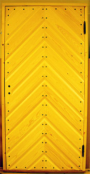 Gul dörr med diagonal panel i fiskbensmönster. Panelbrädorna möts i mitten och skapar ett V-format mönster. Dörren är förstärkt med rader av synliga nitar längs skarvarna, och gångjärnen sitter på höger sida.