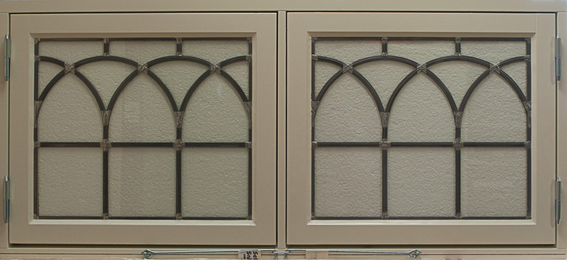 Två sidohängda fönster med beige karm och spröjs i blyinfattat mönster. Glasrutorna är dekorerade med tre spetsbågar i gotisk stil på varje fönster, vilket ger ett klassiskt och hantverksmässigt uttryck. Bakom syns ett frostat eller strukturerat glas som släpper in ljus men skymmer insyn.
