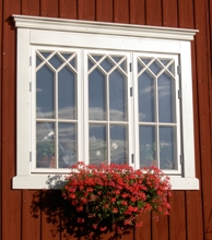 3-luftigt vitmålat spröjsat fönsterparti på röd träfasad med krysspröjs upptill, totalt 27 mindre glasrutor, och blomlåda med röda blommor under fönstret.