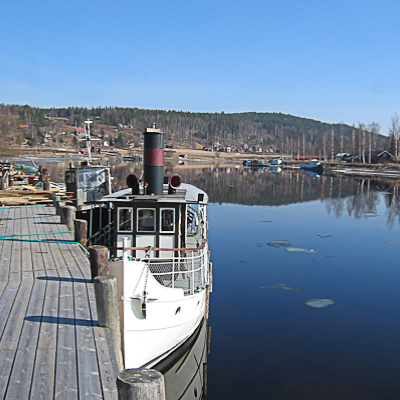 En vit båt ligger förtöjd vid en träkaj en stilla vårdag. Vattnet är spegelblankt och reflekterar båten, skogsklädda kullar och bebyggelse på andra sidan sundet under klar blå himmel.